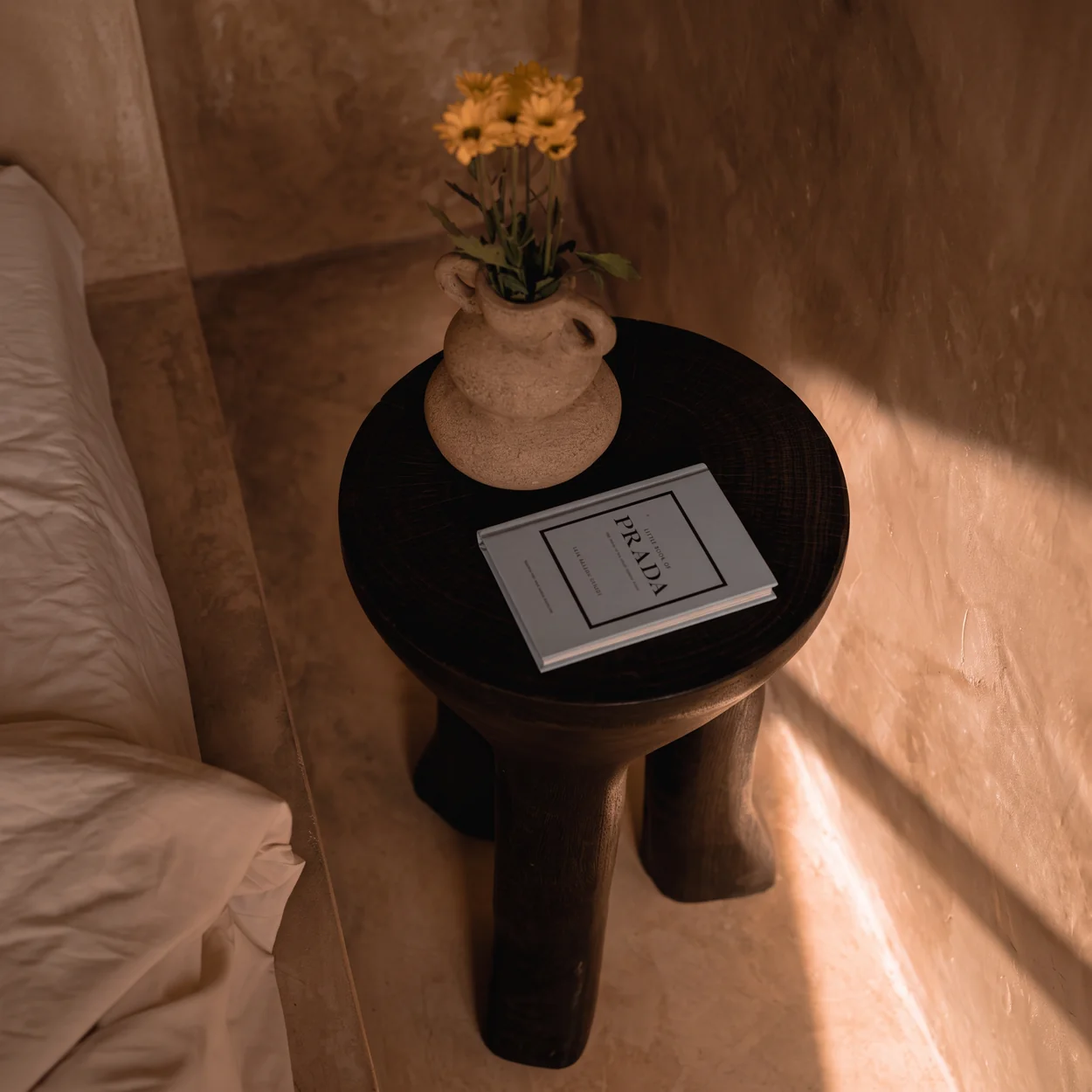 Dark sculptural side table with a ceramic vase, flowers and a book in warm evening light